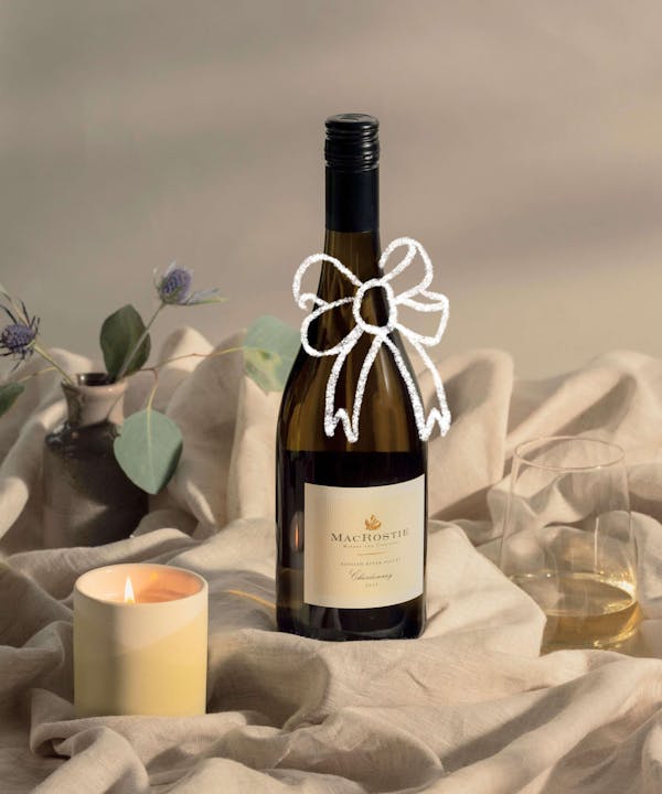 A chardonnay from MacRostie winery and vineyards set on-top of a textured table cloth with a yellow candle to it's left. A textured illustration of a bow is animating on-top of the bottle.