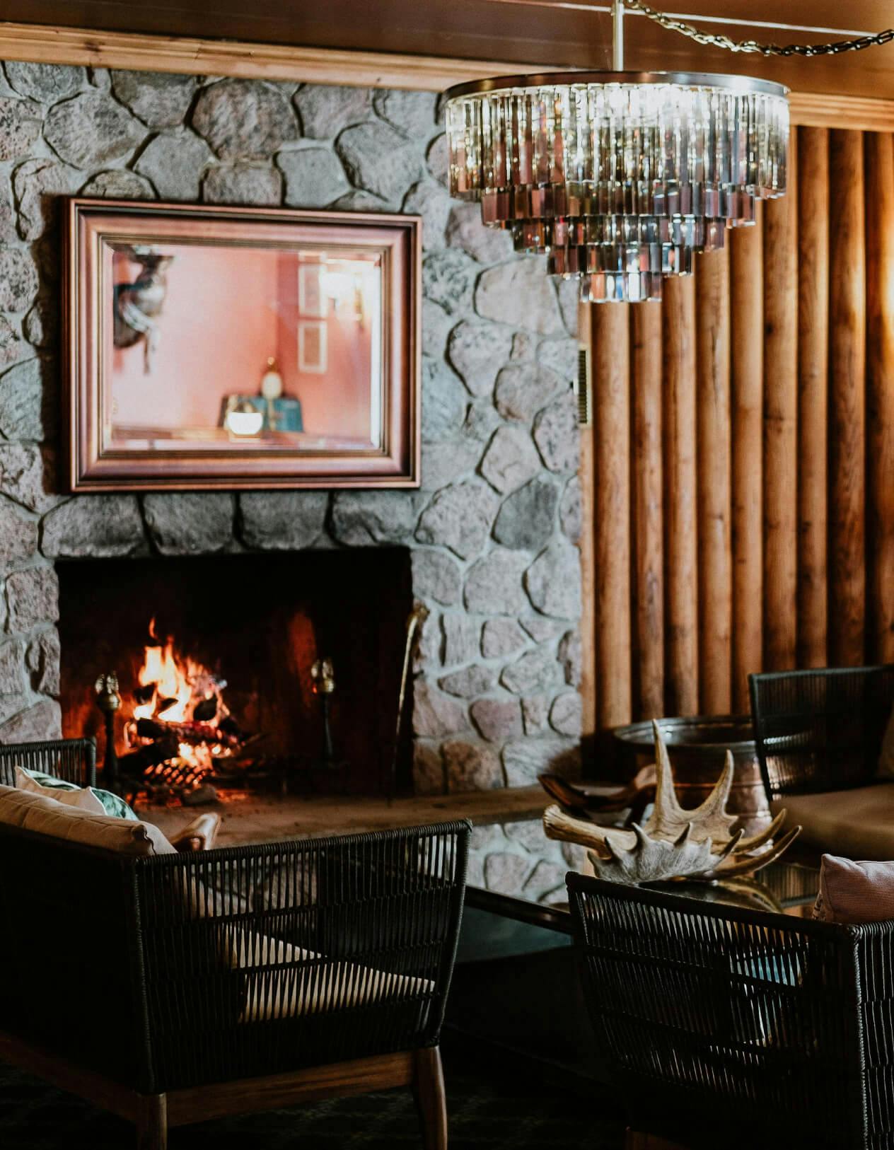 A hotel restaurant with a stone fireplace that has a fire burning within it. There is a mirror above the fireplace reflecting a red wall. Hanging from the ceiling is a mirrored chandelier. In front of the fireplace is a glass coffee table with a single deer antler on top of it. There are black wicker chairs around the table.