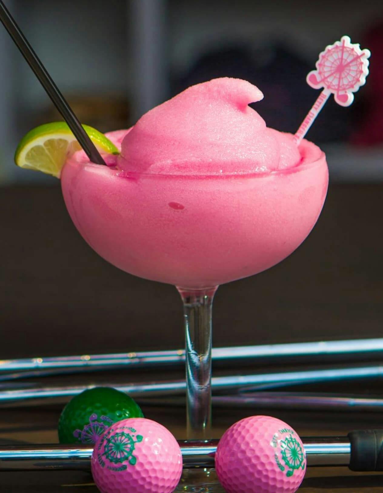 A coup with a pink slushy drink within it garnished with a lime. In front of the drink are two pink golf balls and to the left, there is one green golf ball. Layered across the table the drink is on are multiple golf clubs.