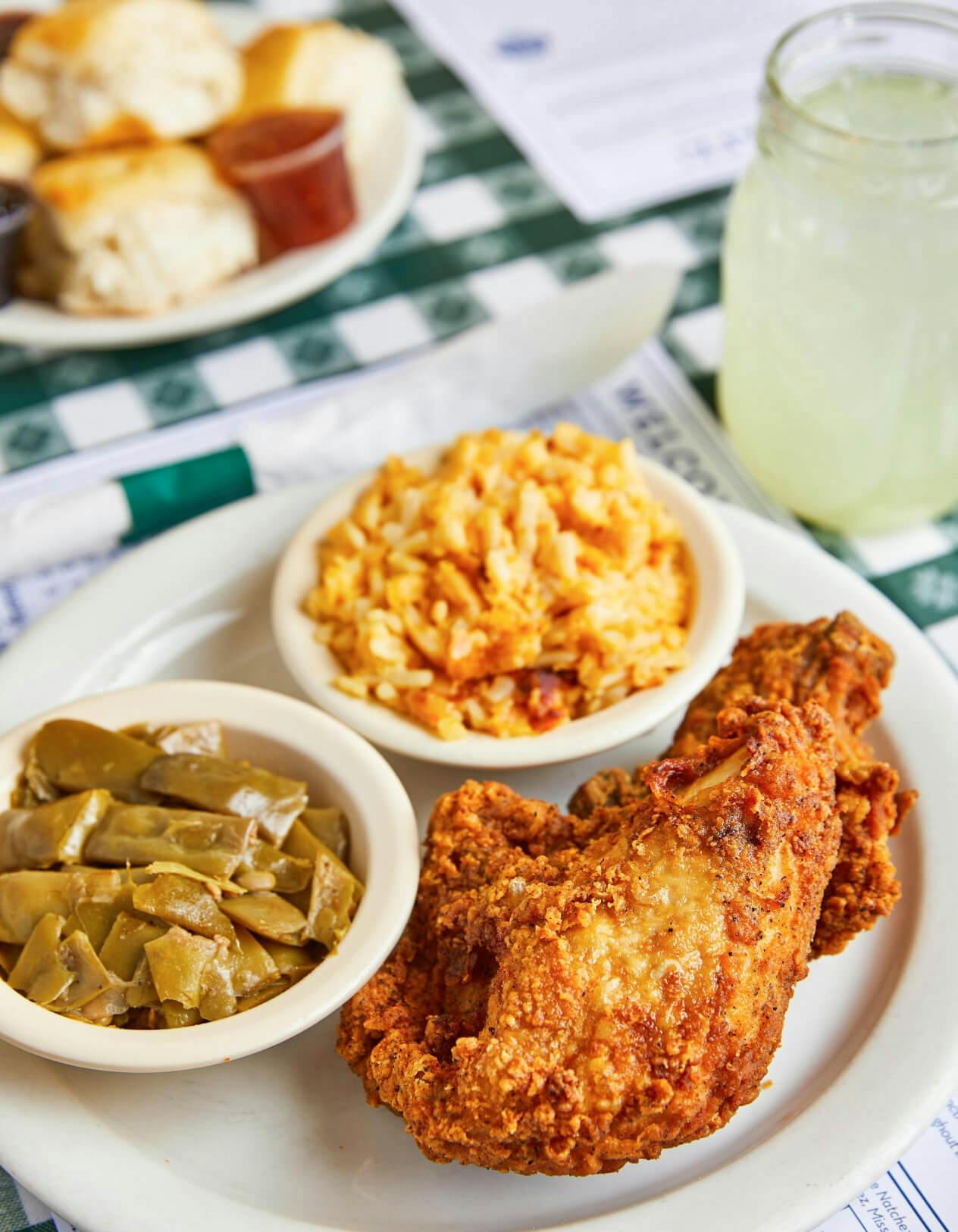 A plate with a piece of fried chicken, green beans, and mac and cheese sits on top of a menu on a green checkered tablecloth. There is a mason jar of lemonade to the right of the plate of food and behind it is another plate with a pile of biscuits that is slightly out of focus.