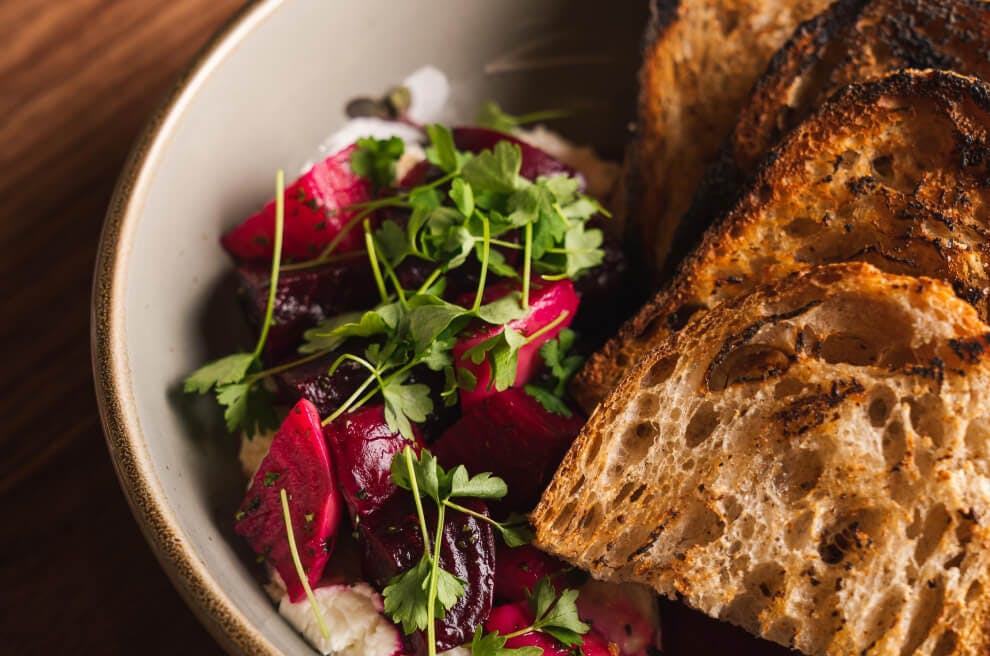 Four pieces of toast sit in a grey bowl. To the left of the toast is a white puree with a pile of bright red beets on top of it that is garnished with green veggies. Everything is on top of a wood grained table.