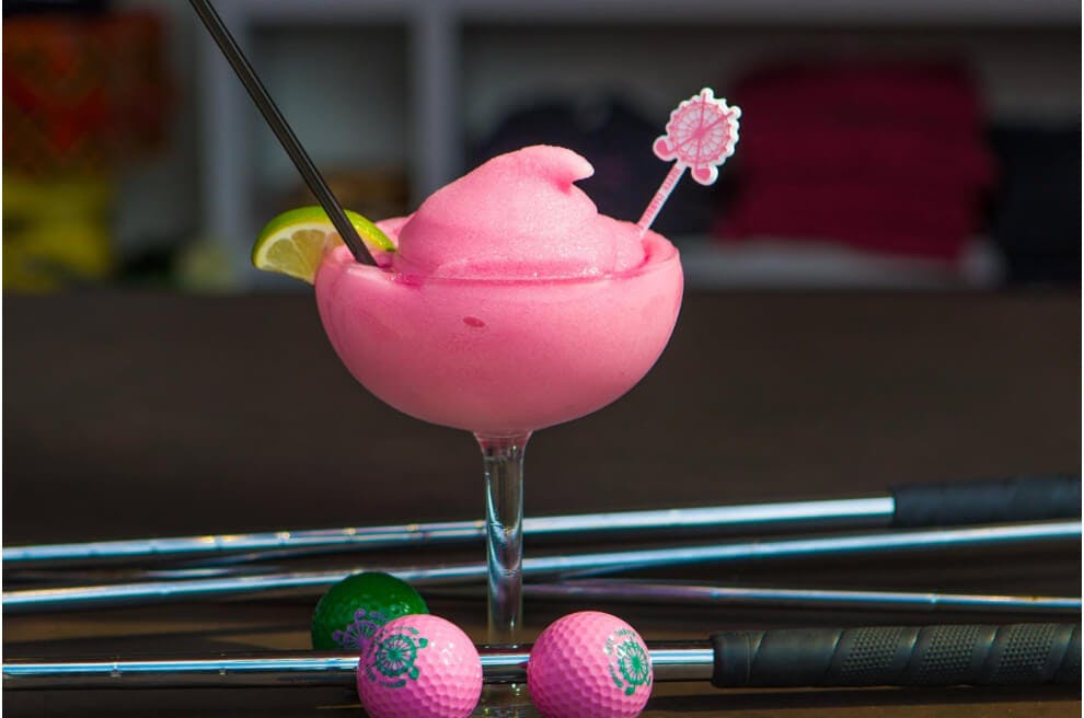 A coup with a pink slushy drink within it garnished with a lime. In front of the drink are two pink golfballs and to the left there is one green golf ball. Layered across the table the drink is on are multiple golf clubs.