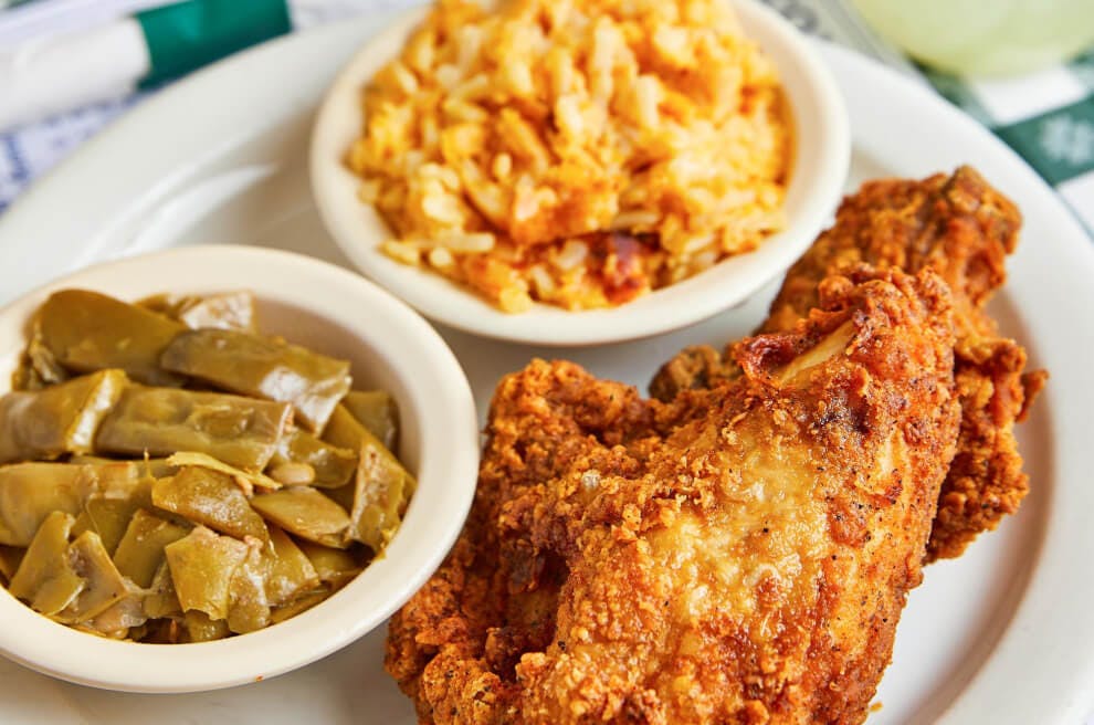 A plate with a piece of fried chicken, green beans, and mac and cheese sits on top of a menu on a green checkered table cloth. There is a mason jar of lemonade to the right of the plate of food and behind it is another plate with a pile of biscuits that is slightly out of focus.