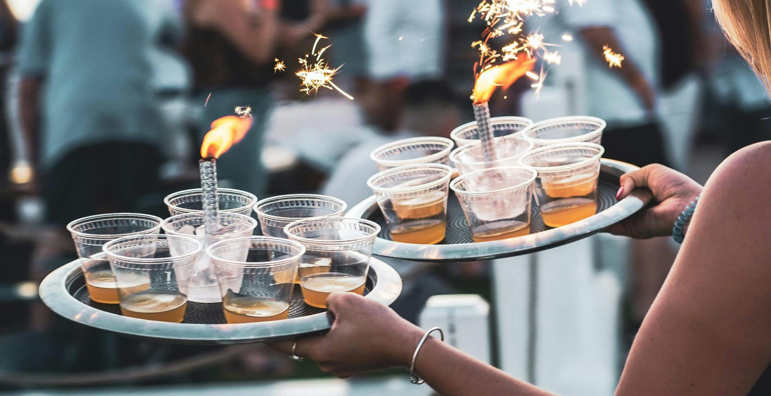 An image of a person carrying two trays of cocktails. Each tray has a lit sparkler in the middle.