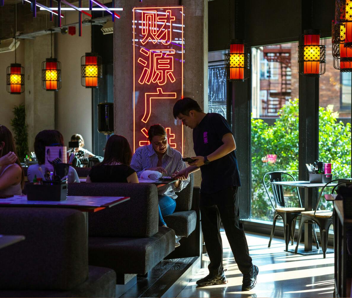 Server interacting with guests at a table in a warmly lit restaurant and a large neon sign in the background with large windows letting in natural light