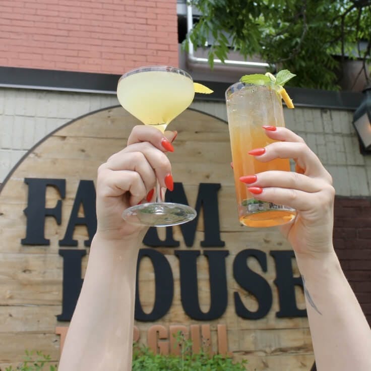 Person holding 2 vibrant-colored cocktails outside of The Farmhouse