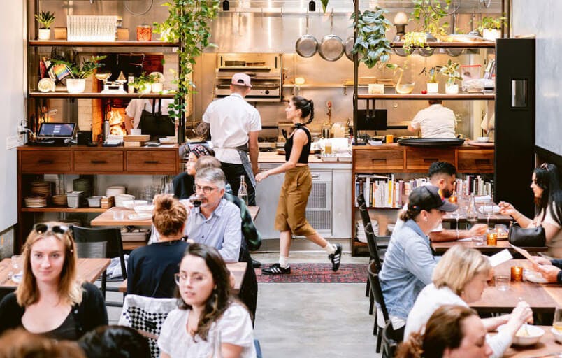 Busy restaurant interior with several diners and servers in the background engaging with an open kitchen at Penny Roma in San Francisco, CA