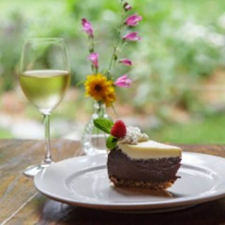 Glass of white wine, cake on a plate, and a vase with flowers on a table outside of Orchard
