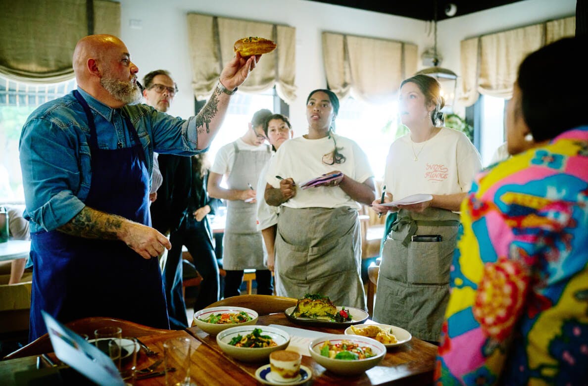Chef's gathering around dishes that will be served to guests in a warmly lit restaurant with large windows letting in natural light