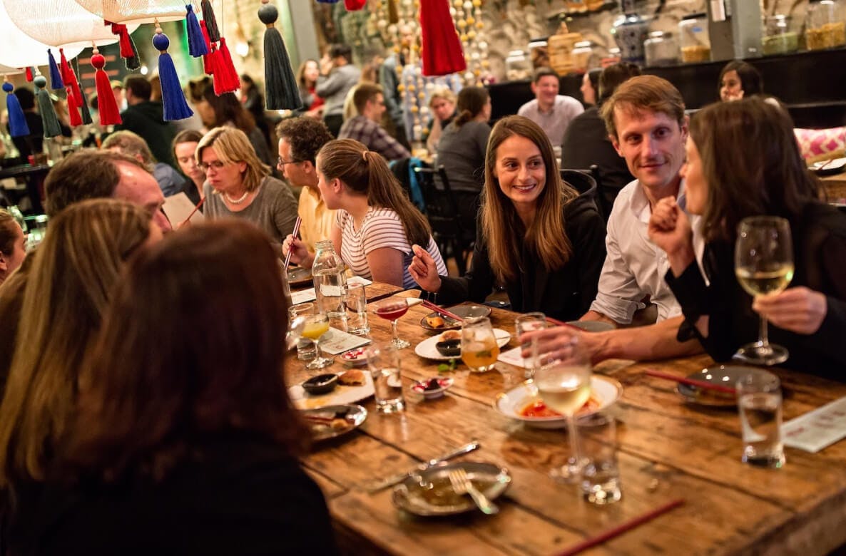Diners gathering around long tables in a warmly lit restaurant hall with wine and small dishes