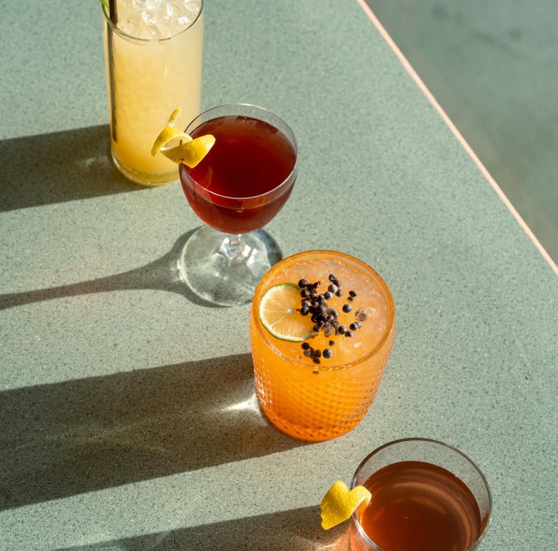An image of four cocktails in a row casting dramatic shadows. In the back of the row is a yellow cocktail in a tall ice-filled glass. In front of that is a deep red cocktail in a coup garnished with a lemon. In front of that is an orange cocktail in a textured rocks glass garnished with a lime and small berries. In the front of the row is an amber cocktail in a rocks glass garnished with a lemon.